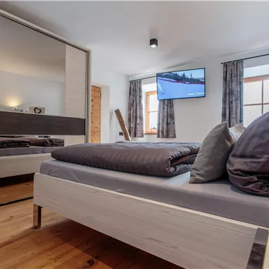 Ein modernes Schlafzimmer mit einem großen Bett, eleganten Kissen und einem Spiegel. Ein Fernseher ist an der Wand montiert, und eine Kerze sorgt für eine gemütliche Atmosphäre.