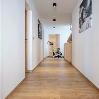 Ein heller Flur mit Holzfußboden und Wänden in Weiß. Am Ende des Flurs steht ein Fitnessgerät und einige Bilder sind an der Wand aufgehängt.