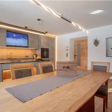 Eine moderne Küche mit Holzdetails und einer großen Esstisch. An der Wand hängt ein Fernseher und die Beleuchtung sorgt für eine angenehme Atmosphäre.