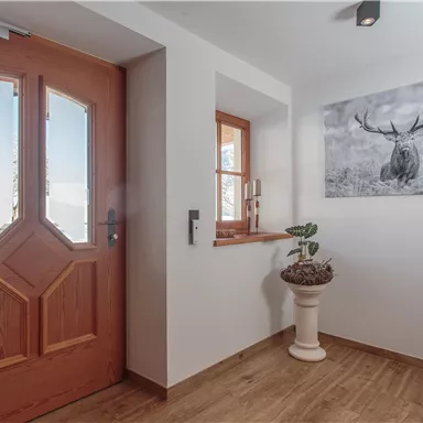 Ein heller Eingangsbereich mit einer Holztür und großen Fenstern. An der Wand hängt ein Bild eines rehbocks, und in der Ecke steht ein Pflanzengefäß.