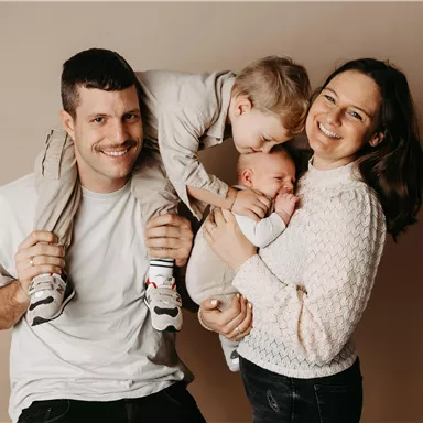 A cheerful family poses together. The father is carrying a child on his shoulders, while the mother holds the baby in her arms.