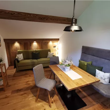 Ein gemütlicher Wohnraum mit einem grünen Sofa und einem Holz-Esstisch. Es gibt eine Kombination aus Stühlen und gemütlichen Sitzplätzen, die eine einladende Atmosphäre schafft.