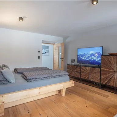 Ein modernes Schlafzimmer mit einem Holzbett, gemütlichen Kissen und einem großen Fernseher. Der Raum hat einen warmen Holzfußboden und eine angenehme, einladende Atmosphäre.