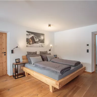 Ein modernes Schlafzimmer mit einem großen Holzbett und grauer Bettwäsche. Der Raum hat helle Wände und warmen Holzboden, sowie zwei Bilder an den Wänden.