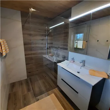 Ein modernes Badezimmer mit einer begehbaren Dusche und eleganten Holz- und Fliesenakzenten. Der Waschbeckenbereich ist mit einem großen Spiegel und minimalistischem Design ausgestattet.