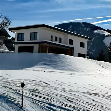 Ein modernes Haus in einer verschneiten Landschaft. Im Hintergrund sind Berge und blauer Himmel zu sehen.