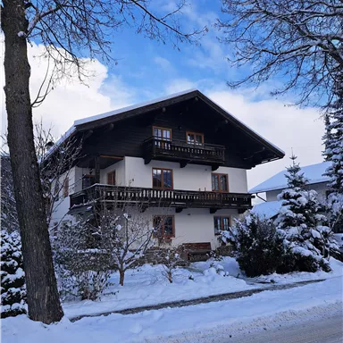 Ein großes, traditionelles Haus im Schnee mit einem blauen Himmel. Umgeben von verschneiten Bäumen und einer ruhigen Straße.