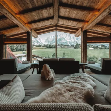 Ein gemütlicher Raum mit Holzdecke und großen Fenstern, die einen Blick auf die Berge und die Natur bieten. Die Einrichtung ist modern und einladend, mit bequemen Sofas und einer warmen Atmosphäre.