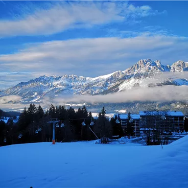 Eine winterliche Landschaft mit schneebedeckten Bergen und einem klaren blauen Himmel. Im Vordergrund liegt eine schneebedeckte Fläche, während Wolken über den Bergen hängen.