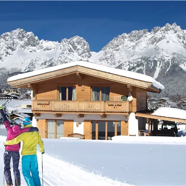 A cozy wooden house in the snow, surrounded by snow-covered mountains. Two skiers are enjoying the winter landscape.