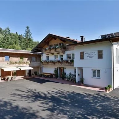 Ein charmantes Gebäude im alpinen Stil mit Balkonen und einer einladenden Fassade. Umgeben von Bäumen und Bergen in klarem blauen Himmel.