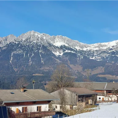 Eine majestätische Berglandschaft mit schneebedeckten Gipfeln und klarem blauem Himmel. In der Vordergrund sind traditionelle, alpine Häuser zu sehen.