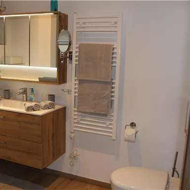 Ein modernes Badezimmer mit einem Holzschrank, einem Spiegel und Handtüchern. Es gibt eine Dusche, eine Toilette und einige dekorative Elemente.