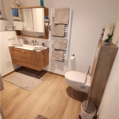 Ein modernes Badezimmer mit einem Holzwaschtisch, einem Spiegel und Handtüchern. Auf dem Boden liegt ein Teppich und die Wände sind in neutralen Tönen gehalten.