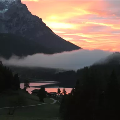 A picturesque mountain landscape with a calm lake and soft clouds. The sky is tinted in warm colors at sunset.