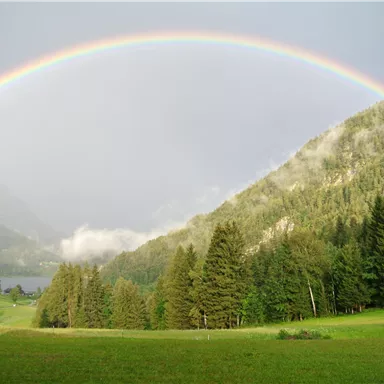 An impressive rainbow spans the mountains. The valley is surrounded by lush forests and a peaceful landscape.