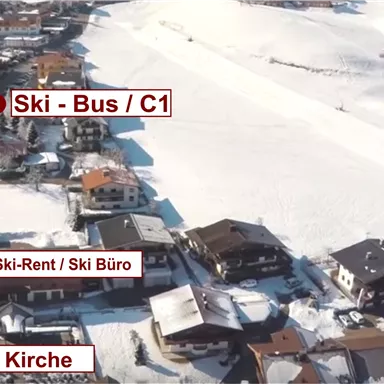 Aerial view of a snow-covered village with labeled locations such as a church, ski bus stop, and rental station. The surroundings appear to be a popular ski area.