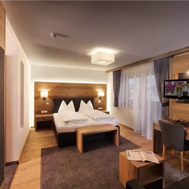 A modern hotel room with wooden details, a double bed, and a small desk. The room is bright and cozy, with a television and windows that provide plenty of daylight.