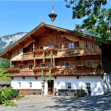 Ein traditionelles Holzhaus mit blumengeschmückten Balkonen. Im Hintergrund sind grüne Berge und blauer Himmel zu sehen.
