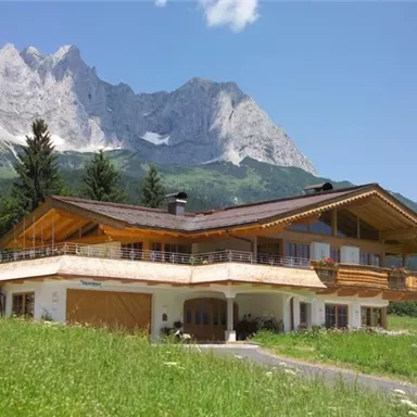 Ein schönes, modernes Chalet mit Holzverkleidung in einer malerischen Berglandschaft. Im Hintergrund sind hohe Berge und ein klarer blauer Himmel zu sehen.