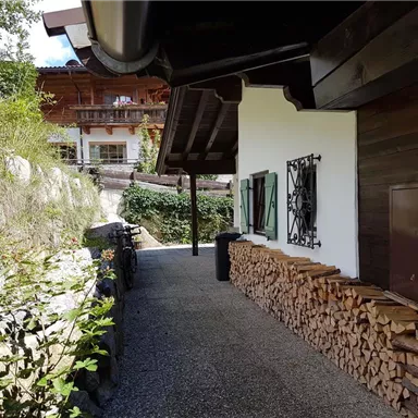 Ein gemütlicher Weg neben einem Holzstapel führt zu einem traditionellen Haus. Umgeben von Grün und Sonne vermittelt die Szene ein rustikales, einladendes Gefühl.