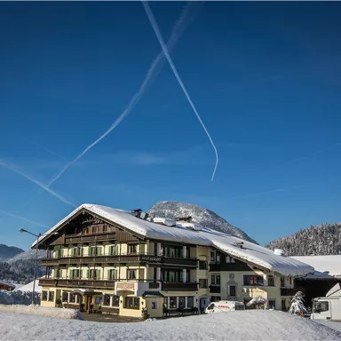 A charming, snow-covered building in the mountains under a clear blue sky. The wooden-clad facade and surrounding winter landscape create a cozy atmosphere.