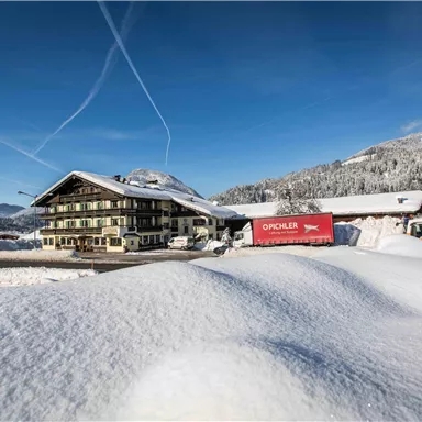 A snow-covered landscape picture with a cozy building and mountains in the background. The sky is clear and bright blue.