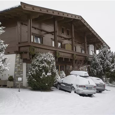 Ein gemütliches Chalet im Winter, umgeben von Schnee. Parkende Autos und schneebedeckte Bäume verleihen der Szene eine ruhige Atmosphäre.