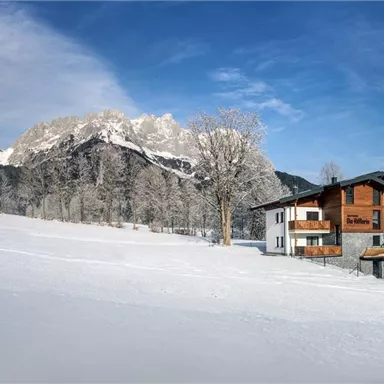 A charming house in a snow-covered landscape with majestic mountains in the background. The trees are frosty and the sky is clear and blue.
