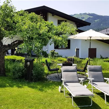 An inviting garden with loungers and a sun umbrella in front of a modern house. In the background, gentle hills and green trees are visible.