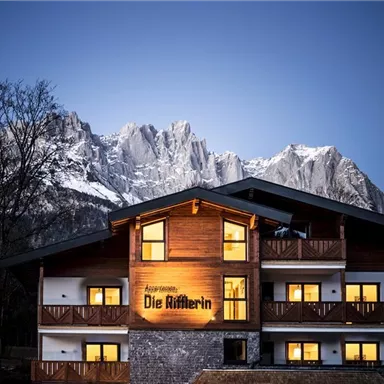 A charming wooden house with warmly lit windows. In the background, majestic mountains rise under a clear sky.