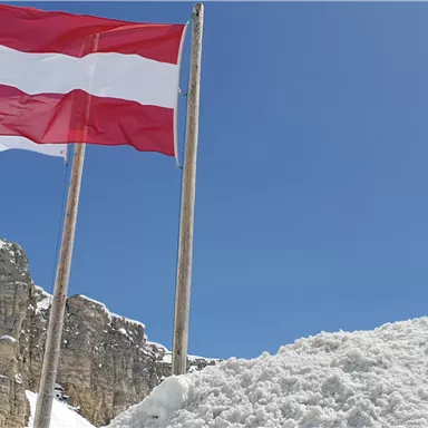 An Austrian flag is waving in front of a clear blue sky. In the foreground, a large pile of snow can be seen.
