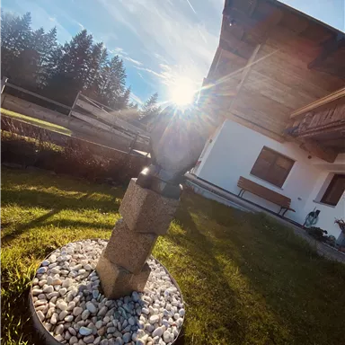 A beautiful garden with a wooden house in the background. In the center stands a decorative sculpture on pebbles, while the sun shines in the background.