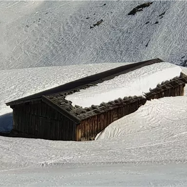 An old wooden cabin, partially covered in snow. It stands in a snowy landscape with gentle hills.