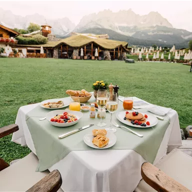 A beautifully set table outdoors with various breakfast dishes and drinks. In the background, green meadows and mountains can be seen.