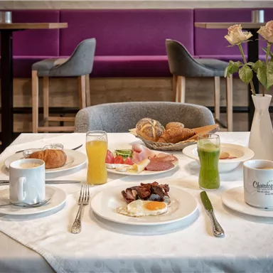 A beautifully set breakfast table with various dishes, including eggs, cold cuts, and rolls. Beverages such as coffee, juice, and a green smoothie are served alongside.