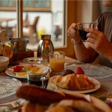 Eine gemütliche Frühstücksszene mit verschiedenen Speisen auf dem Tisch. Eine Person hält eine Tasse Kaffee und genießt eine Auswahl an Früchten und Gebäck.