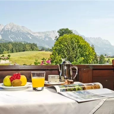 A beautiful breakfast table with fresh fruit, a glass of juice, and a teapot. In the background, the impressive mountains and the green landscape stretch out.