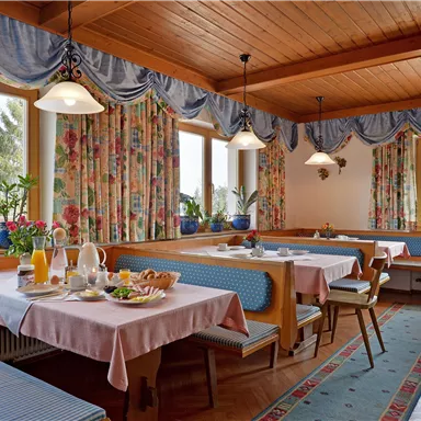 Ein gemütlicher Speisesaal mit Holzmöbeln und hellen Tischdecken. Die Fenster sind mit floralem Vorhang geschmückt und es sind Pflanzen auf den Tischen.