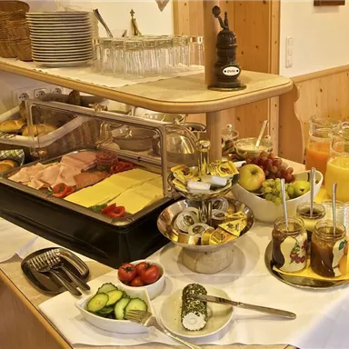Ein Frühstücksbuffet mit verschiedenen Wurst- und Käsesorten, frischem Obst und Gemüse. Zusätzlich gibt es Getränke wie Saft in Glaskaraffen.