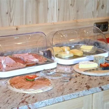 Ein Buffet mit verschiedenen Wurst- und Käsesorten, die unter Kunststoffhauben präsentiert werden. Neben dem Buffet befinden sich Teller und Besteck auf einem Holzpanel.