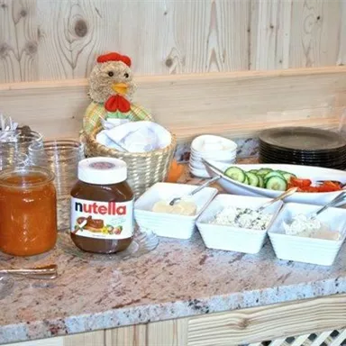 Ein Frühstückstisch mit verschiedenen Marmeladen, Nutella und belegten Tellern. Frisches Gemüse wie Gurken und Paprika sind ebenfalls neben einer dekorativen Figur zu sehen.