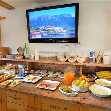 Ein Buffet mit verschiedenen Frühstücksgerichten und Getränken. Im Hintergrund ist ein Fernseher mit Landschaftsbild.