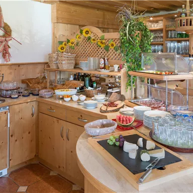 Ein gemütlicher Frühstückstisch mit verschiedenen Holz- und Steinplatten, reichlich Sattigkeiten wie Käse, Aufschnitt und frischem Obst. Die warme Holzführung und die Dekoration sorgen für eine einladende Atmosphäre.