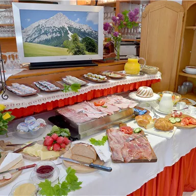 Ein reichhaltiges Frühstücksbuffet mit verschiedenen Fleisch- und Käseplatten, frischem Obst und Brötchen. Im Hintergrund ist ein Bild von Bergen zu sehen.