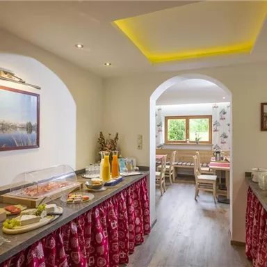 Ein einladender Frühstücksraum mit einem Buffet voller Leckereien. Im Hintergrund sieht man gemütliche Sitzplätze und Fenster mit Blick ins Grüne.