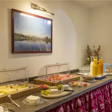 Ein Frühstücksbuffet mit verschiedenen Käsesorten, Aufschnitt und Obst. Im Hintergrund sieht man orange Saft und ein hübsches Bild an der Wand.