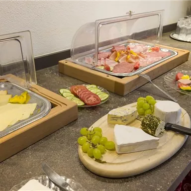 Ein reichhaltiges Buffet mit verschiedenen Käsesorten und Aufschnitt. Neben Trauben sind auch frisches Obst und Säfte zu sehen.