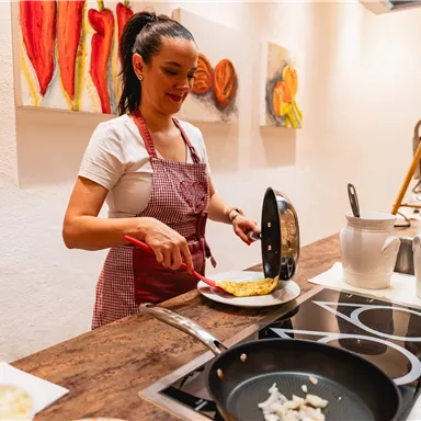 Eine Frau in einer Küche bereitet ein Omelett zu. Sie trägt eine Schürze und steht am Herd.