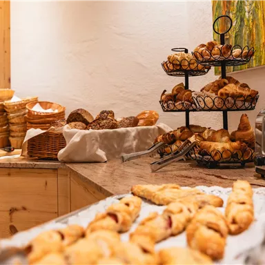 Ein gemütlicher Frühstückstisch mit verschiedenen Gebäcksorten und Brötchen. Im Hintergrund stehen Körbe mit frischem Brot und ein Toaster.